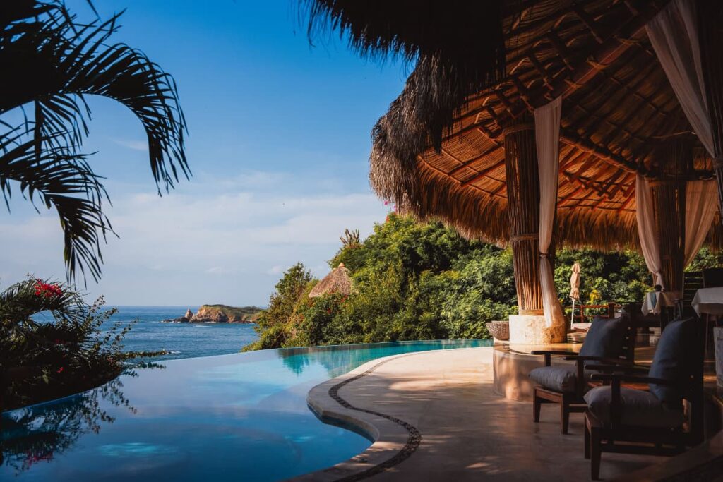 A view of infinity pool at ZOA resort featuring a thatched roof, comfortable chairs, lush green hills and a partial ocean view.