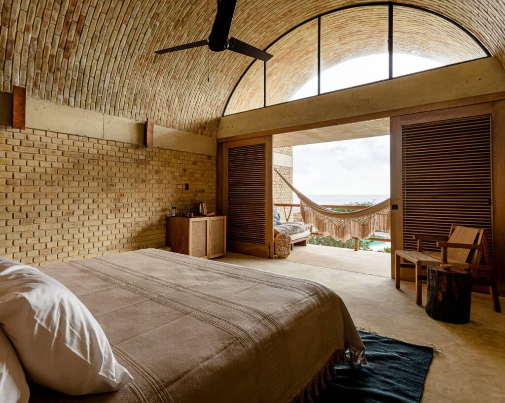 A minimalistic bedroom at Casa Sforza furnished with a comfortable bed, a cabinet, a chair, an arched roof with a ceiling fan with a hammock in the private space outdoor.