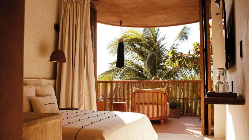 A view of the room at Xique Hotel furnished with a bed, a TV and private outdoor space with a chair surrounded by palm trees.