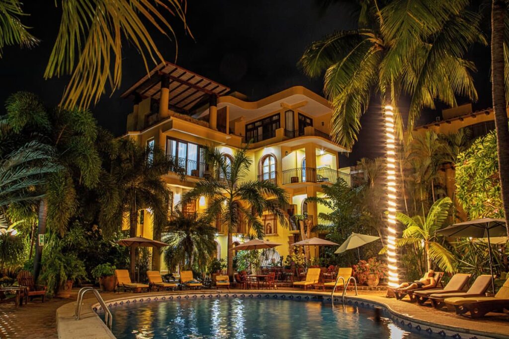 A view of the outdoor pool at Suites La Hacienda surrounded by palm trees and lush greenery and the hotel property.