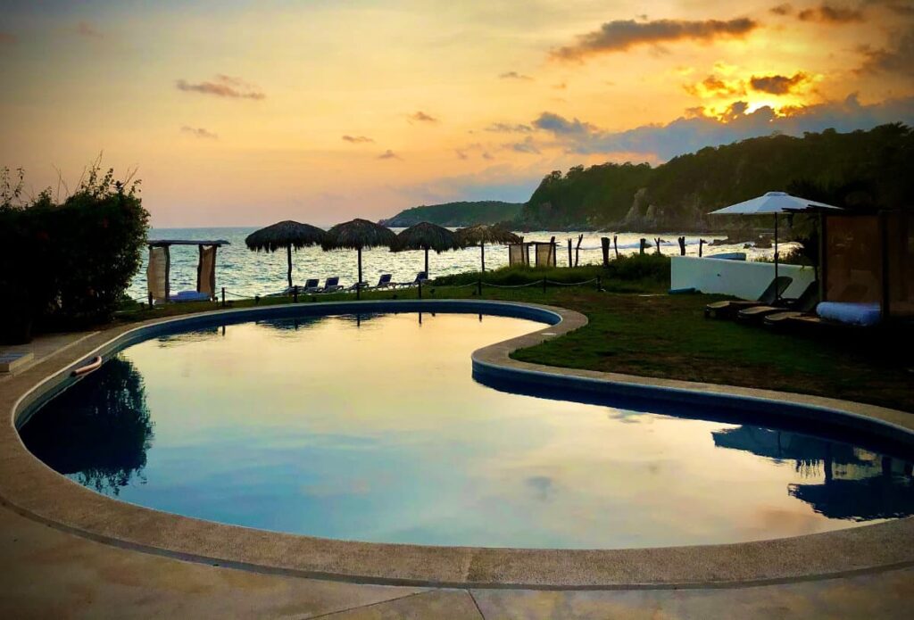 A view of sunset from Manta Raya Hotel with the reflection of sky in the swimming pool, thatched parsols with sunloungers overlooking the ocean and lush green hills.