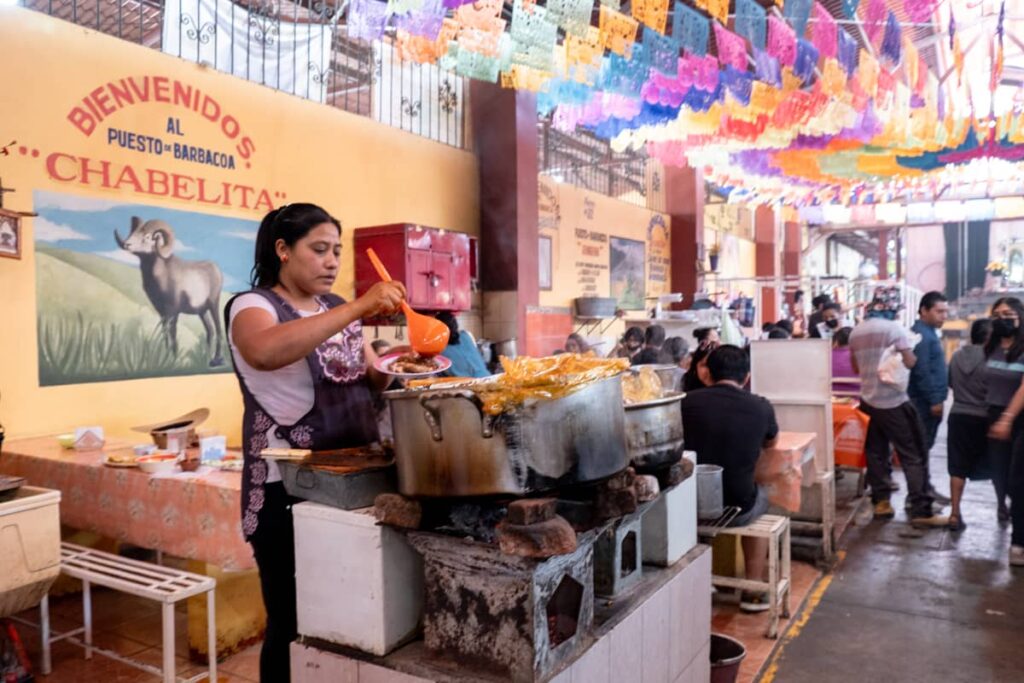 A woman ladles consome from a large pot of barbacoa into a bowl at the Tlacolula Sunday Market. Behind her is a sign painted on the wall that reads, Biendvenidos al Puesto de Barbacoa Chabelita.