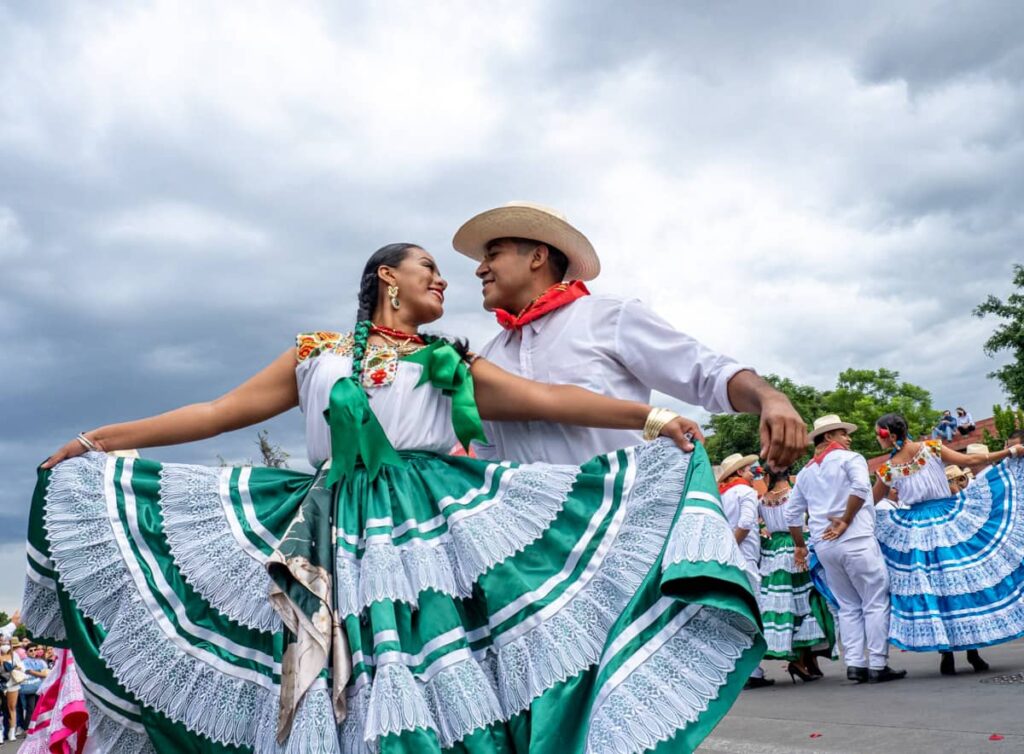 At the Guelaguetza Festival in Oaxaca, a woman wearing a green and white dress with green ribbons in her hair dances with a man in white wearing a hat and red scarf around her neck.