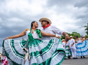 At the Guelaguetza Festival in Oaxaca, a woman wearing a green and white dress with green ribbons in her hair dances with a man in white wearing a hat and red scarf around her neck.