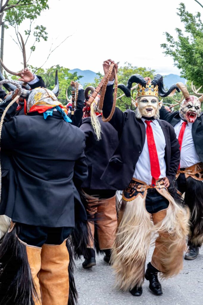 Men wearing a suit jacket, red tie, long blonde fur chaps, and devil masks with curly horns gather and dance before the start of a Guelaguetza parade. In their hands are braided leather whips.