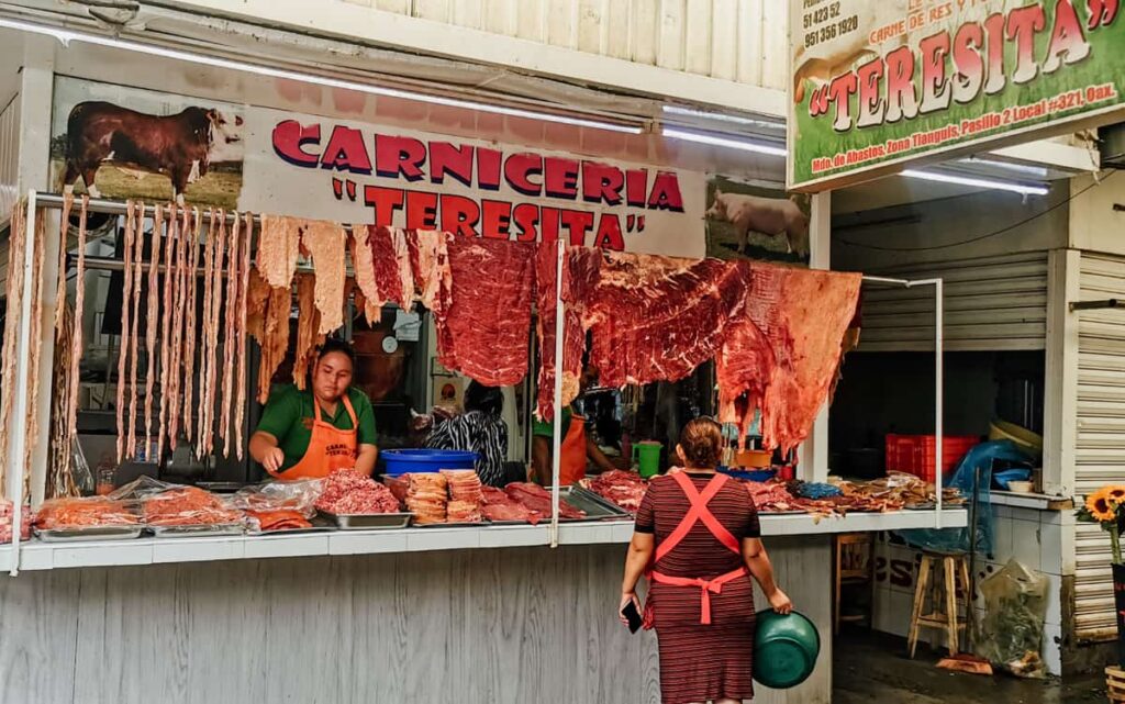 A woman wearing an orange apron arranges the display at a meat stall inside Abastos Market in Oaxaca. On the counter are stacks of various meat cuts. Above her head hangs tasajo and other thin cuts to dry.