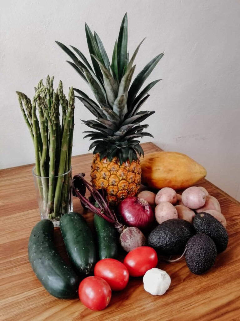 Fresh produce from a Oaxaca market sits on a wooden table. The pile includes a papaya, pineapple, cucumbers, avocados, garlic, potatoes, a red onion, beet, and asaparagus.