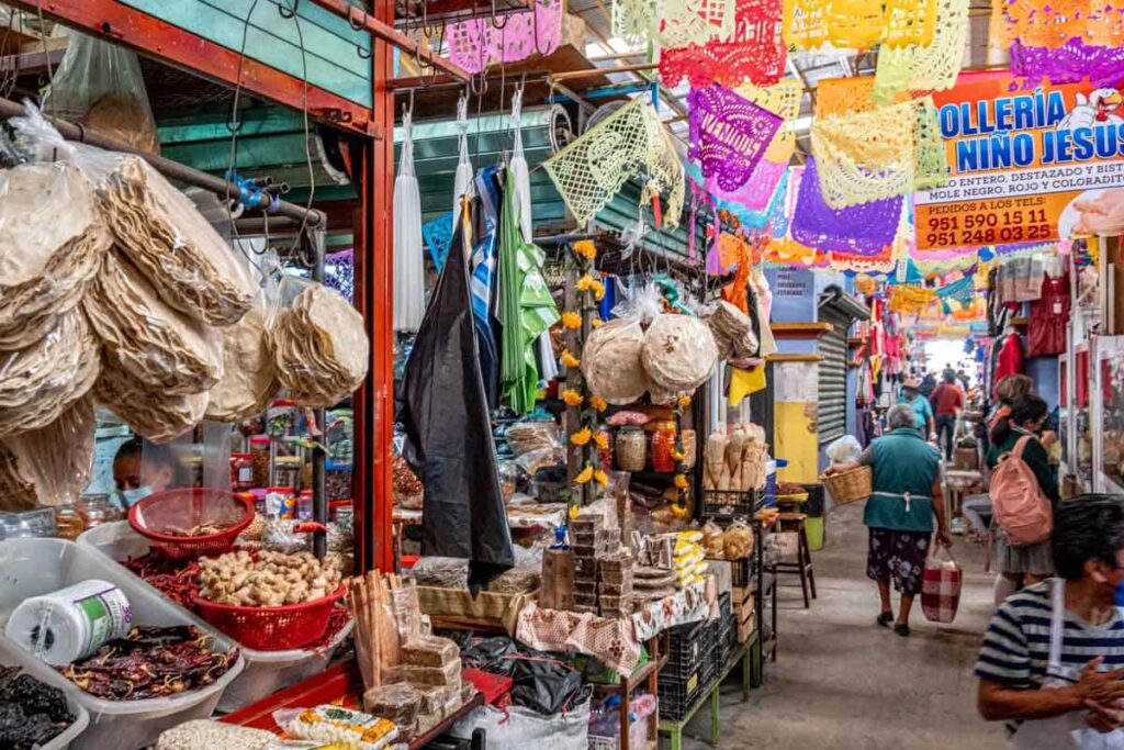 People walk through the aisles of the Ocotlán Market which are full of products - dried chiles and tortillas. Above hang colorful papel picado decorations.