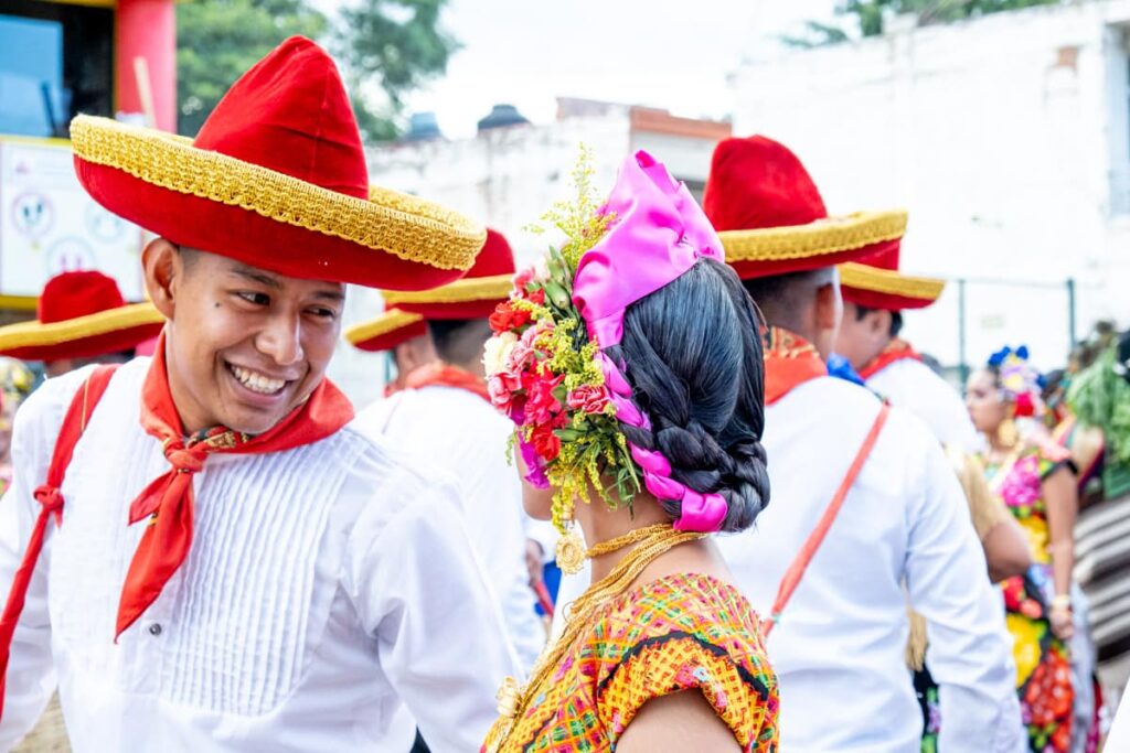 A man wearing a white shirt with a red handkerchief around his neck and a red velvet hat with gold trim smiles at a women in a colorful top with pink ribbon and flowers in her hair.