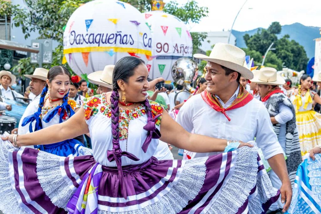 At the Guelaguetza in Oaxaca, a woman fans her dark purple and white skirt as she smiles at her dance partner. Her hair is braided and woven with purple ribbon. Behind them are other dancers and large white spheres made of fabric that are common in parades.