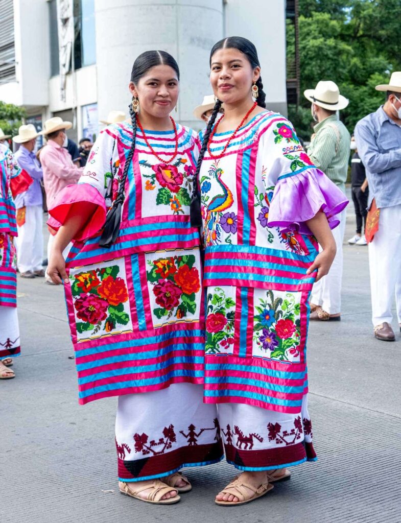 Two women from La Canada region of Oaxaca pose for a picture during the Guelaguetza Festival. They are dressed in traditional clothes featuring flower and bird prints with predominantly pink and blue colors. Their dark hair is braided into thin pigtails.