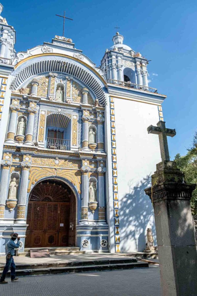 A man wearing a cowboy hat walks past the baby blue and gold exterior of the church in Ocotlán Oaxaca.
