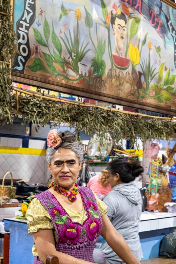 In Ocotlán Oaxaca, Beatrice, aka Frida, poses for a picture wearing a colorful apron, necklace, and flowers in her hair.