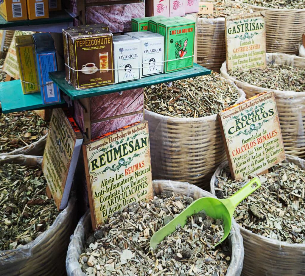 Woven baskets of herb blends for sale at the Abastos Market in Oaxaca with signs listing the name and benefits of each. On a shelf above, various herbal blends are sold in boxes.