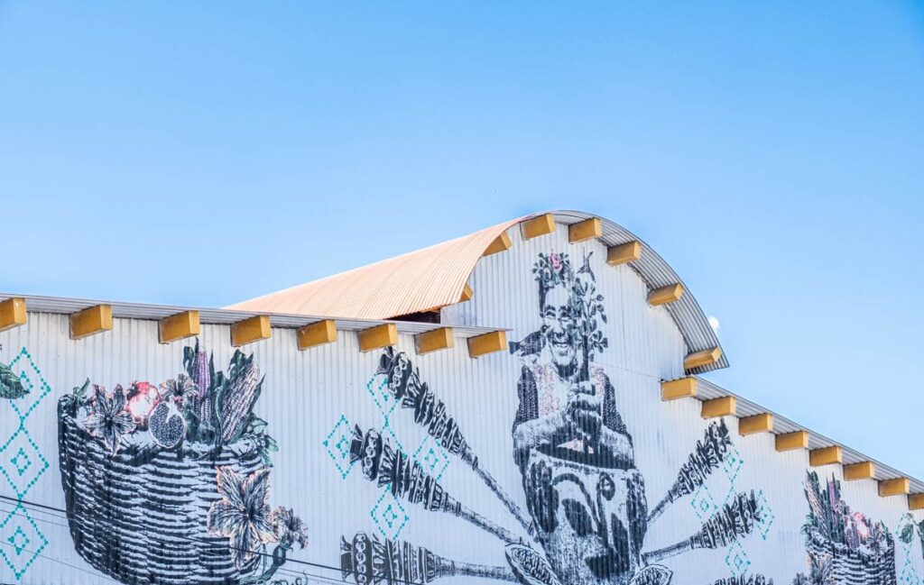 The signage of the Ocotlán Market features a black and white graphic mural that depicts a woman frothing a pot of drinking chocolate with baskets of fresh fruits and vegetables on either side of her.