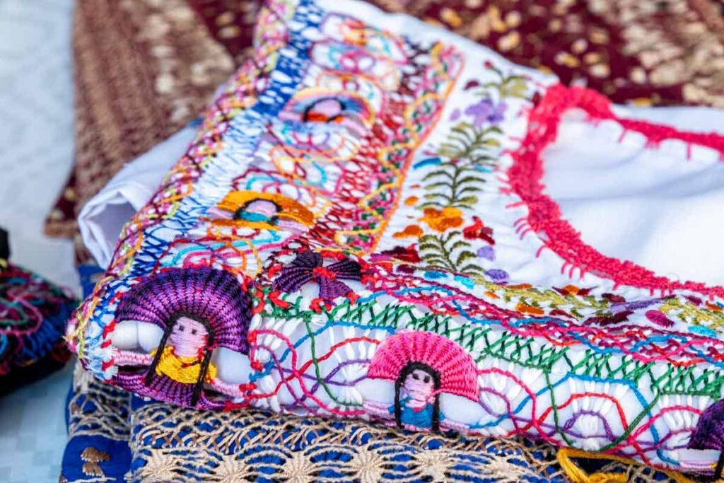 Folded blouses with colorful woven threads depicting flowers, figures, and other designs are stacked on a table at the Ocotlan market in Oaxaca