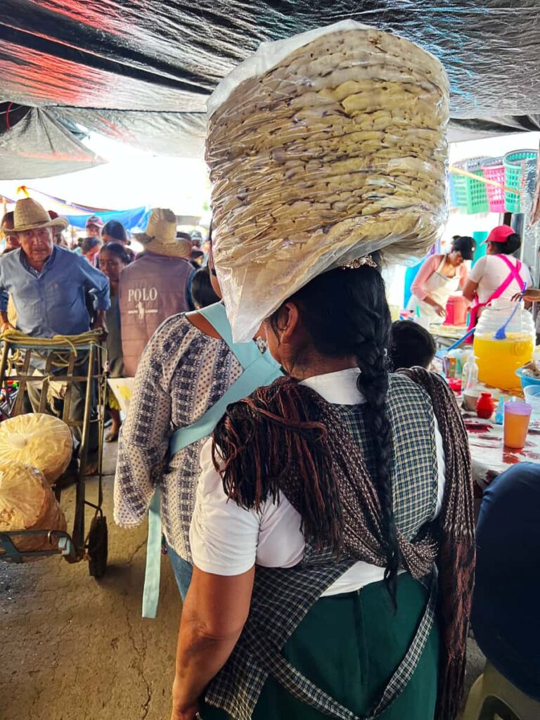 A woman walks through Abastos Market in Oaxaca with a stack of tlayuda tortillas wrapped in a plastic bag on top of her head. The back view shows a long black braid down her back with a checker print apron and a green skirt.
