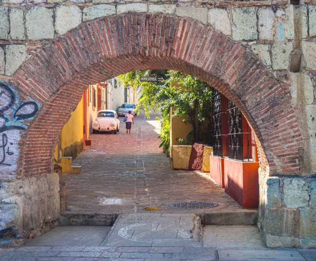 A woman walks next to a white VW bug parked along an alley in Xochimilco Oaxaca, framed by the stone and brick archway of the aqueduct.