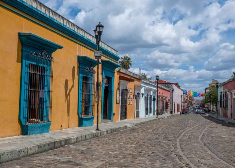A cobblestone street in Centro Oaxaca Mexico with colorful buildings. In the distance are colorful flags and some cars.