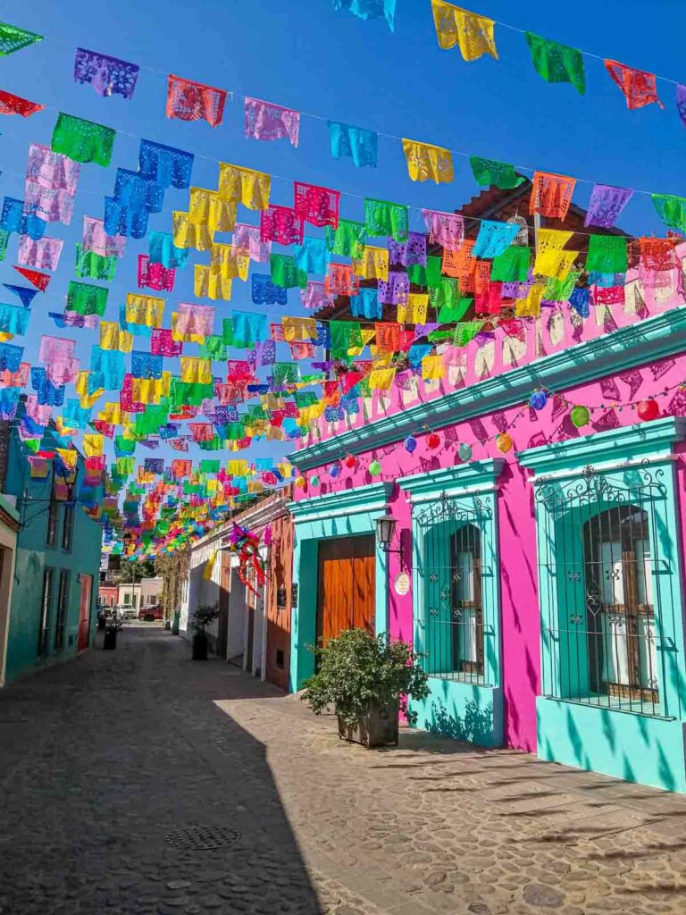 Colorful flags called papel picado hang over the cobblestone street in Jalatlaco neighborhood. The street is lined with colorful buildings, in particular one that is bright pink with aqua trim.