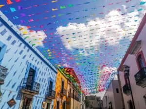 A street in Oaxaca Centro looking up shows colorful papel picado flags and buildings in a range of colors with small iron balconies.