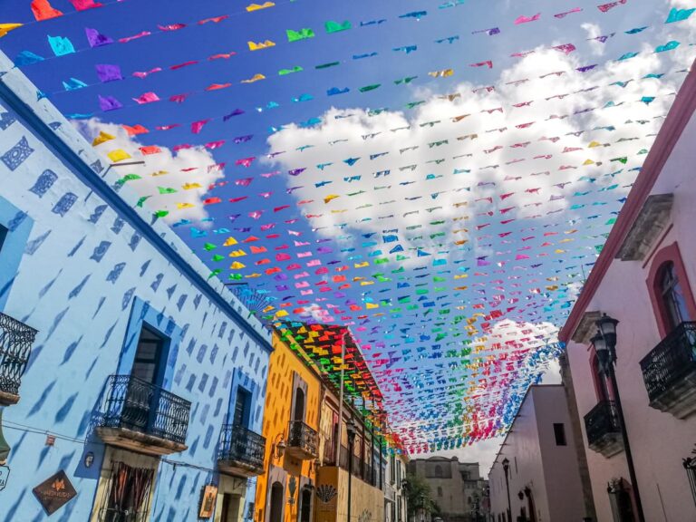 A street in Oaxaca Centro looking up shows colorful papel picado flags and buildings in a range of colors with small iron balconies.