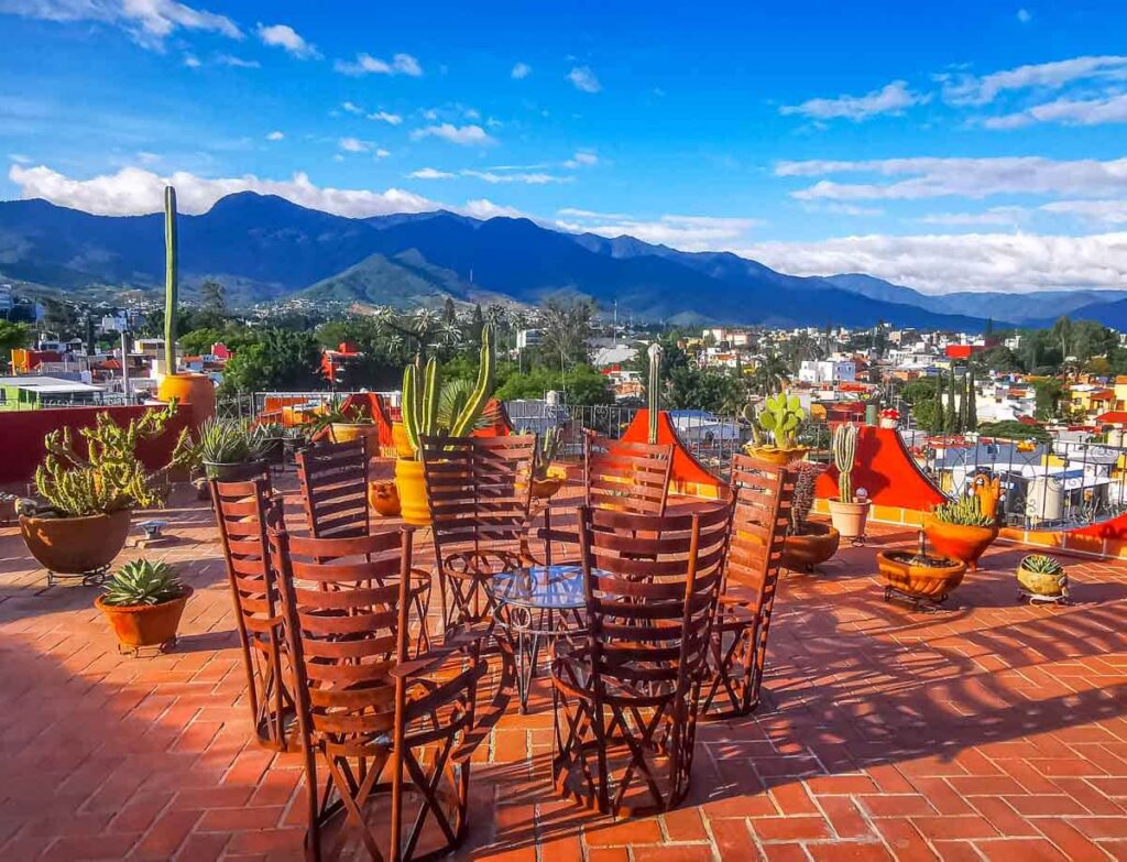 A rooftop mountain view from Xochimilco Oaxaca. The paved terrace is full of cactus and other plants as well as a metal table and chairs. In the background are buildings and mountains.