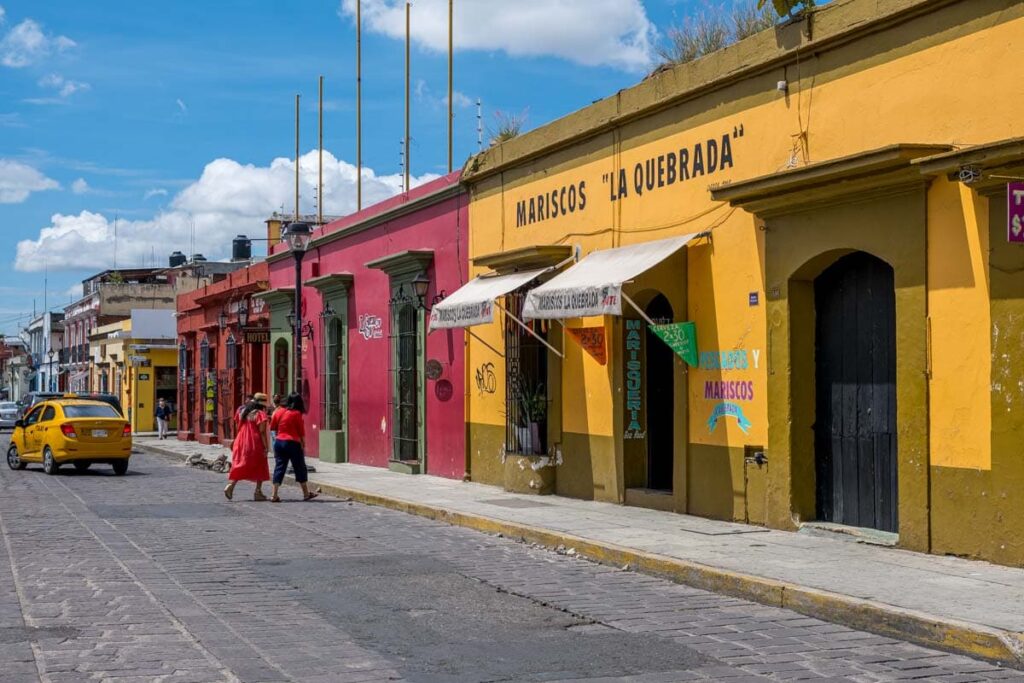 Two women cross the street in Centro Oaxaca Mexico. The buildings on the side are painted a bright yellow, magenta, and deep red.