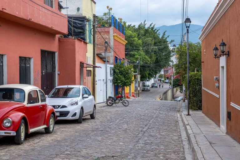 A quiet street in Xochimilco neighborhood, Oaxaca. Several cars and a motorcycle are parked while two people walk in the distance.
