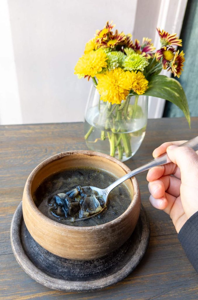 Close-up of a hand holding a spoonful of huitlacoche soup in a rustic clay bowl, at Coeva restaurants in Oaxaca. On the table is a vase of fresh yellow and maroon flowers in the background.