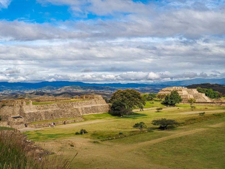 Wide view of the main plaza at Monte Alban in Oaxaca showing stone platforms and pyramids on both sides, green hillsides, and mountains under a cloudy sky.