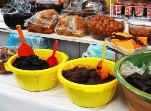 At a market, two yellow plastic bins are filled with Oaxacan mole to buy and cook at home. On the shelf behind the moles are fruits preserved in sugar.