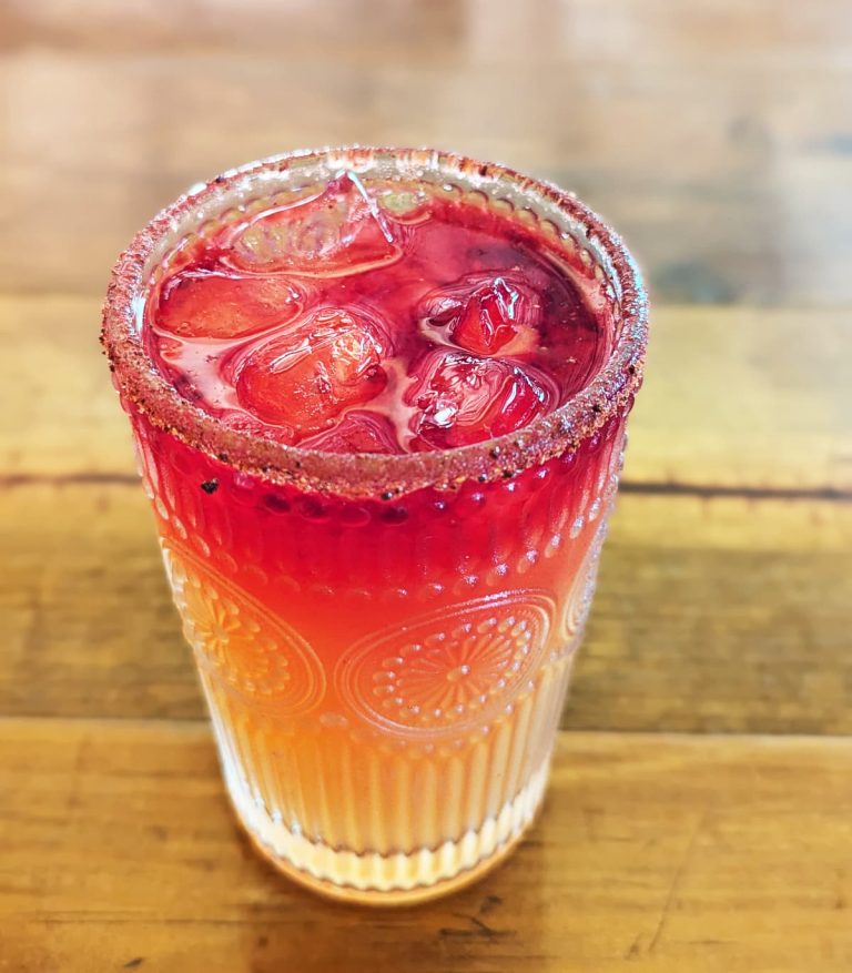 A sugar-rimmed glass of iced tepache, one of the most popular Oaxacan drinks, on a wooden table. The drink is layered with a red color on top to a yellow color on the bottom.