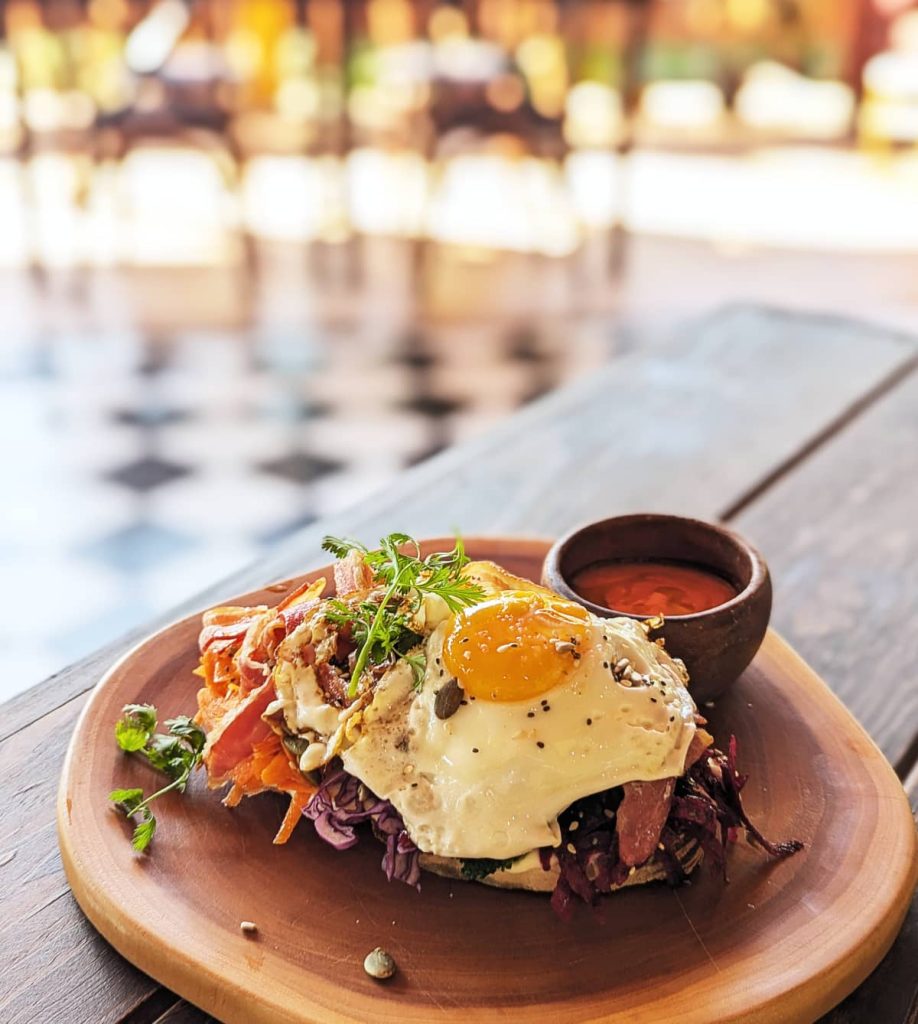 A fried egg, bacon, red cabbage, and herbs on avocado toast served on a wooden plate with a small bowl of gazpacho at Sahumerio, one of the best places to eat brunch in La Punta.