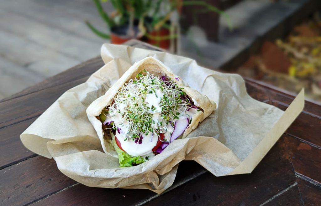 At Cultivo vegan restaurant in Puerto Escondido, a pita stuffed with tempeh, veggies, sprouts, and a white cream sauce sits on a piece of brown wax paper on a wooden table.