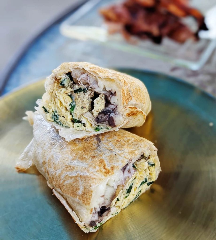 Breakfast burrito of scrambled eggs, beans, spinach, and melted cheese inside a crispy grilled tortilla, with a side of crispy bacon in the background at Disfruta in Puerto Escondido.