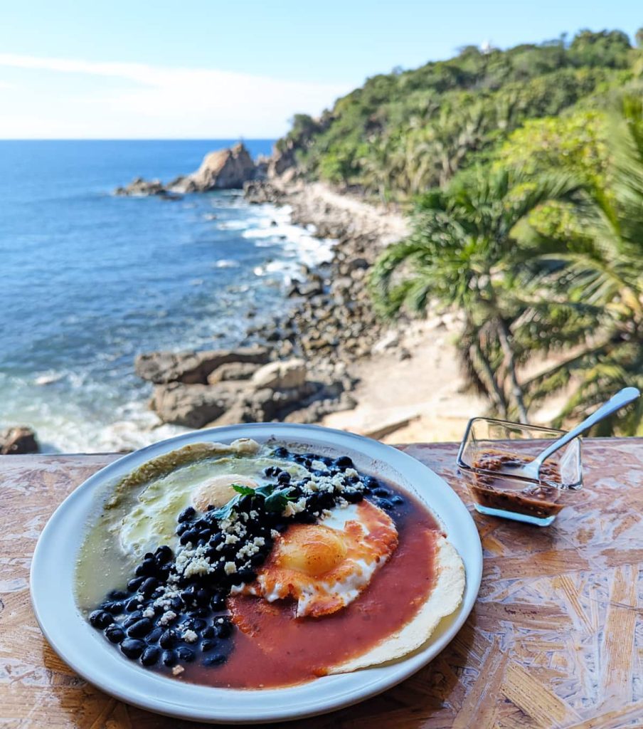 Huevos rancheros with fried eggs, red and green salsa, black beans, and queso fresco on a corn tortilla at Zikaru, with a rocky coastline and palm trees in the background.