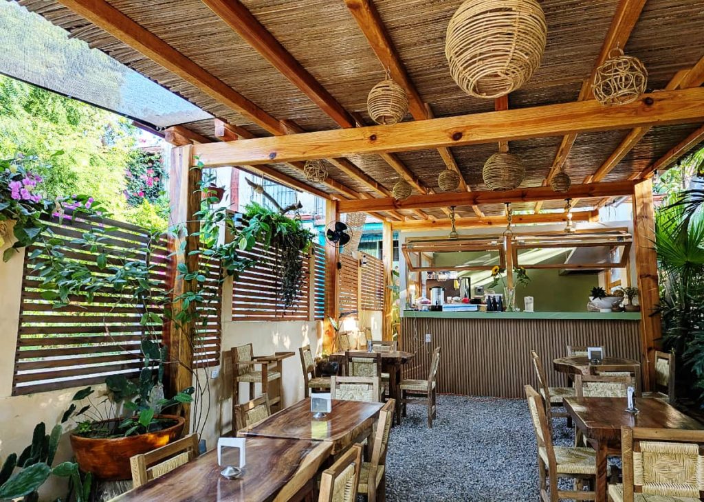 Open-air dining area at Mongo Brunch in Puerto Escondido with wooden tables, woven chairs, rattan pendant lights, potted plants, and a bamboo-roofed palapa over a gravel floor.