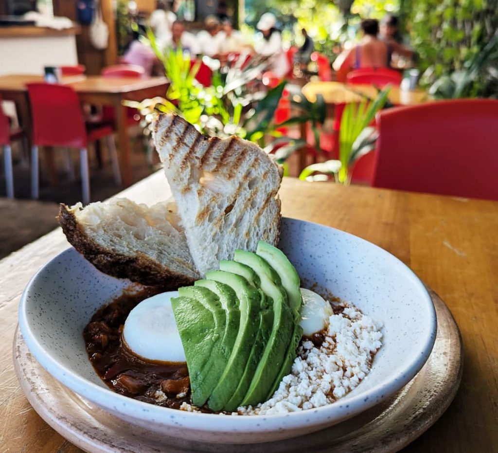 Shakshuka with two poached eggs, sliced avocado, crumbled queso fresco, and grilled sourdough bread in a speckled ceramic bowl at Loloh Panadería, a brunch spot in Puerto Escondido.