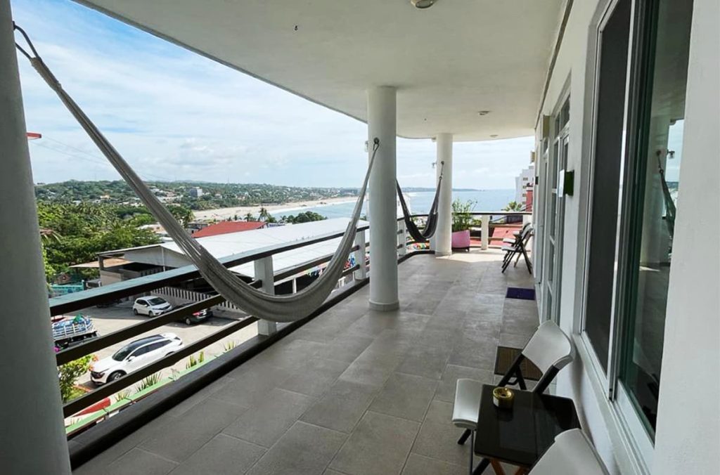 View from the balcony of Casa Mia Hotel in Puerto Escondido looking towards Zicatela Beach. On the balcony are several folding chairs and a small folding table. Two hammocks hang between the columns before the balcony curve to the right.