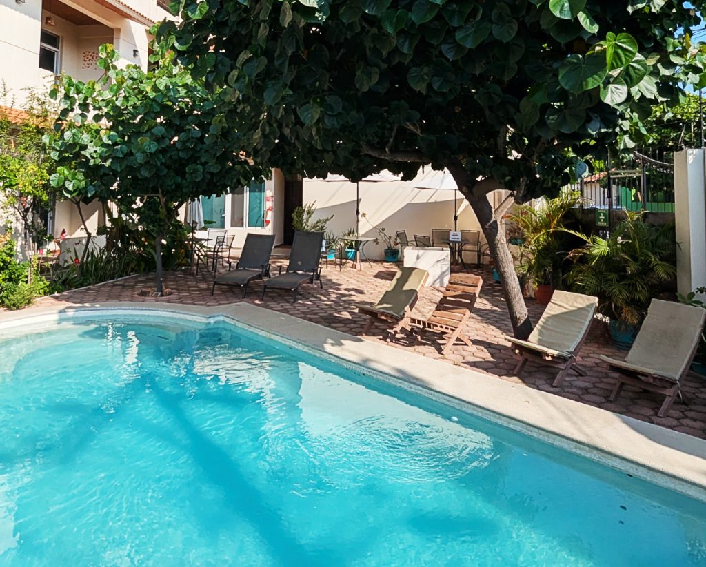 At Casa Puerto Bonito coliving in Puerto Escondido, the tuquoise oval shaped pool is surrounded by folding wooden chairs under a short tree with large dark green leaves. In the back are two loungers under the shade.