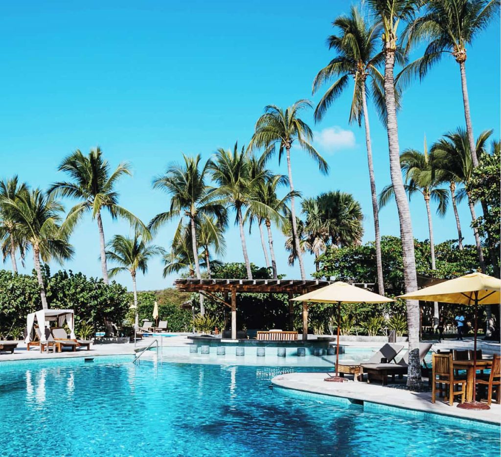 Bright turquoise pool surrounded by tall palm trees at Hotel Suites Villasol in Bacocho. Yellow umbrellas are at one edge of the pool. In the background is a bar with cement stools in shallow water and a cabana with white fabric hanging in all four corners.