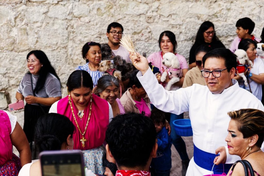 Wearing a white robe with a blue sash, the priest of La Merced church blesses the animals with a natural brush and a bucket of water. People holding their animals surround him.