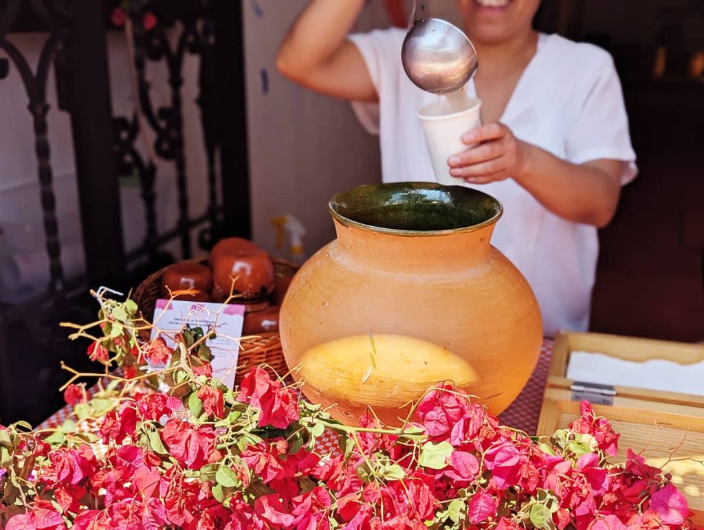 A smiling woman uses a metal ladle to pour a white drink in a paper cup from a brown clay vessel with a green glazed interior. On the table in front are pink bougainvillea flowers.