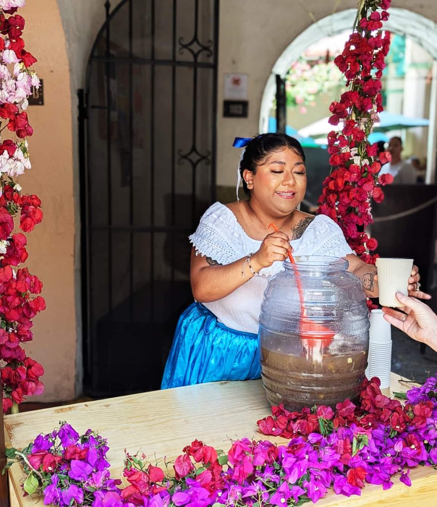 A woman wearing a blue skirt, white blouse, and a blue ribbon in her hair serves a traditional drink, agua de chilacayota, from a tall plastic container using an orange ladle. She is surrounded by pink, red, and magenta bougainvillea flowers.