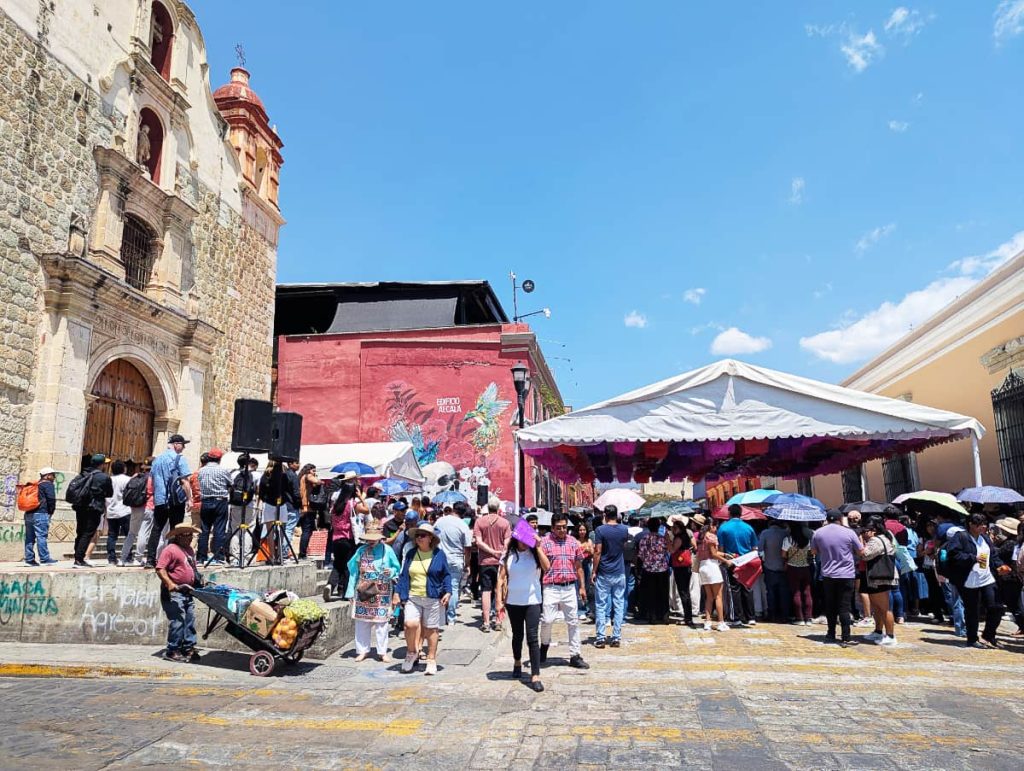 In front of a stone church, a crowd of people gather under a tent in the midday sun for the official Día de la Samaritana festival in Oaxaca.