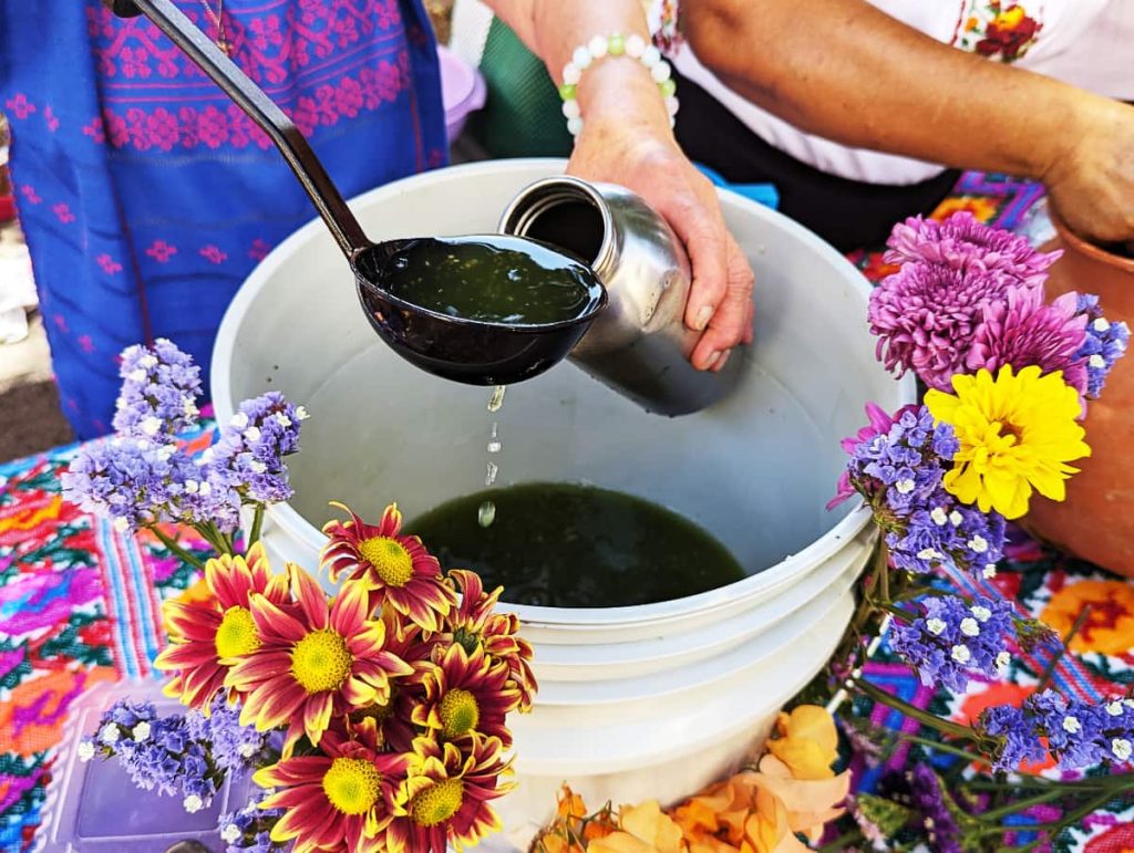 During the Good Samaritan holiday in Oaxaca, a woman uses a ladle to pour agua fresca into a reusable metal bottle from a white bucket. Flowers are attached to the bucket for decoration.