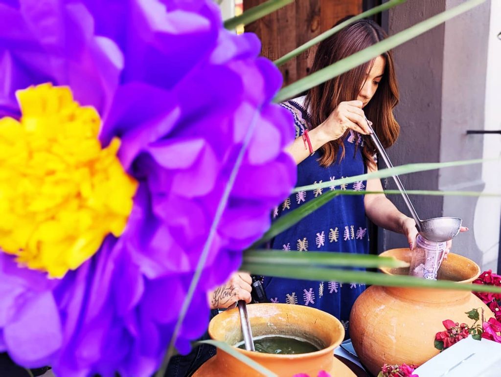 A woman dressed in a traditional blue blouse ladles a drink from a clay pot into a plastic cup during Samaritan's Day in Oaxaca. In the foreground is a large purple flower with a yellow center made of paper.