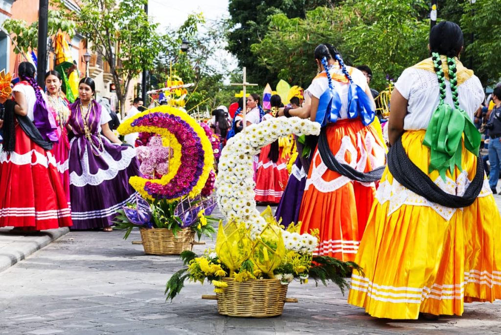 Chinas Oaxaqueñas dressed in traditional skirts, matching scarf, and white shirt stand in the street before a parade in Oaxaca. Their hair is braided long with brightly colored ribbons. And two baskets with large flower decorations rest on the street next to them.