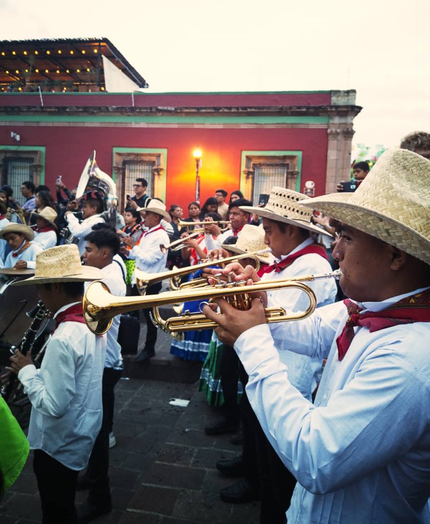 A line of men playing the trumpet in the street during a parade in Oaxaca City wearing a white shirt with a red handkerchief around their necks and a palm hat.